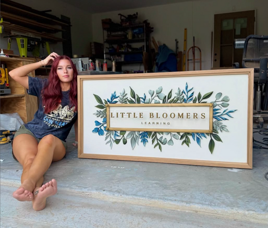 Little Bloomers sign with a white background, natural wood frame, blue and green foliage leaf border, golden inner frame with Marissa sitting next to it in the garage